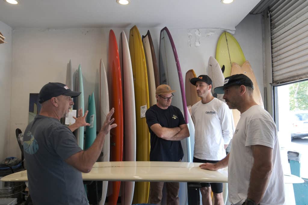Shapers-Club- Quatre hommes discutant des planches de surf Gordon Smith dans un magasin de surf. -surfshop-surfboard