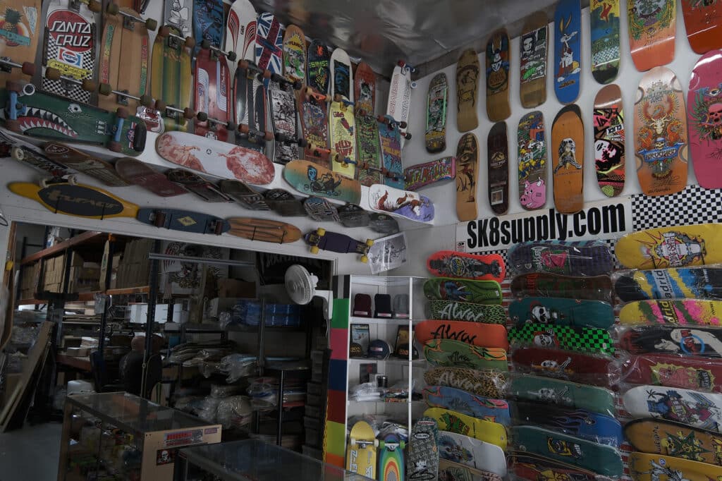 Shapers-Club- Une collection diversifiée de planches de skateboard et de surf colorées exposées sur un mur avec une signalisation « sk8supply.com ». -surfshop-surfboard