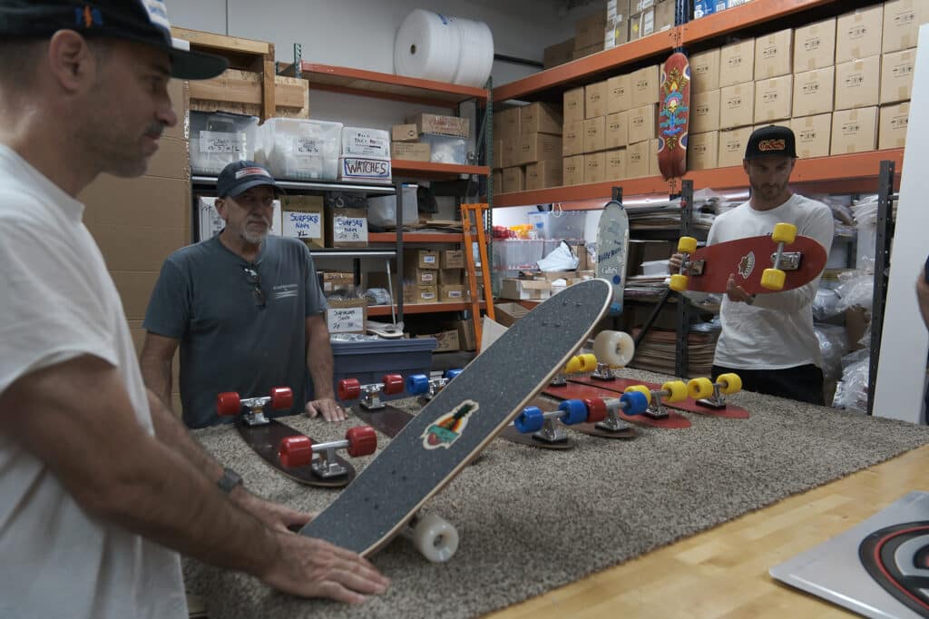 Shapers-Club- Des hommes examinant une planche à roulettes Gordon Smith dans un entrepôt. -surfshop-surfboard