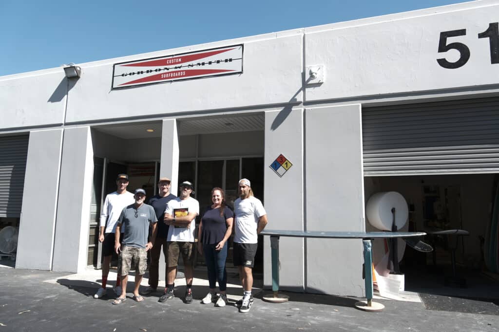 Shapers-Club- Groupe d'individus posant ensemble devant un bâtiment industriel avec une pancarte indiquant « Gordon and Smith Custom Surf. -surfshop-surfboard