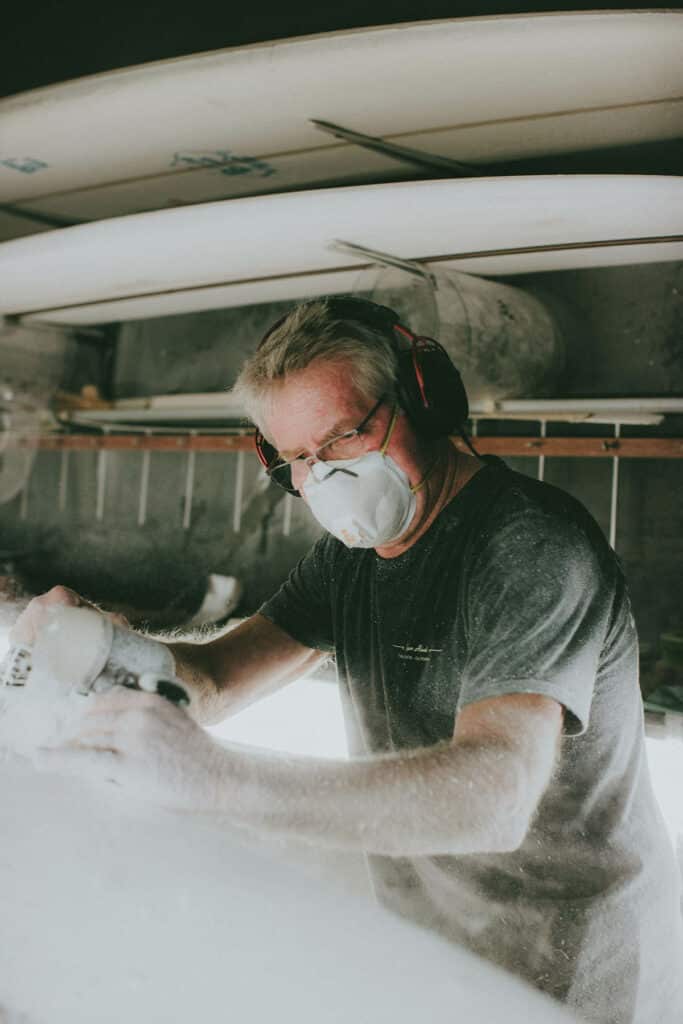 Shapers-Club- Roger Hinds, portant un masque anti-poussière et une protection auditive, façonne une planche de surf dans un atelier, avec des particules de poussière visibles dans l'air. -surfshop-surfboard