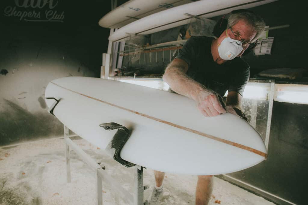 Shapers-Club- Roger Hinds, portant un masque anti-poussière et des lunettes, façonne une planche de surf dans l'atelier de l'Univers du Shape avec des planches de surf en arrière-plan. Des particules de poussière sont visibles dans l'air. -surfshop-surfboard