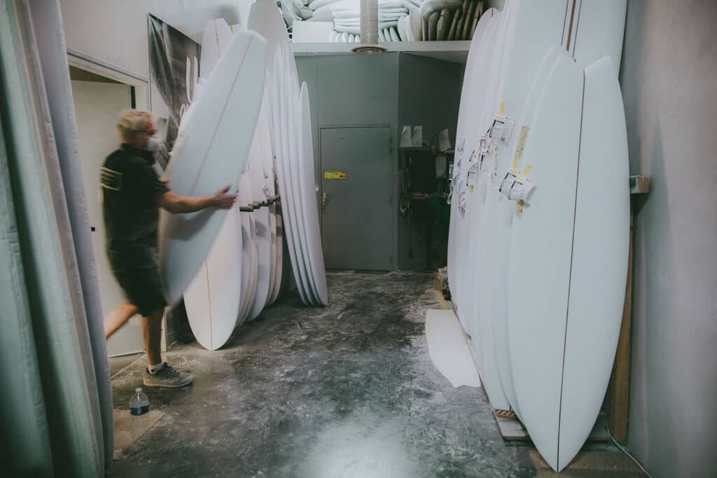 Shapers-Club- Une personne traverse un atelier, l'Univers du Shape, où plusieurs planches de surf fabriquées par le Maître Artisan des Surfboards Roger Hinds sont rangées debout contre le mur. -surfshop-surfboard