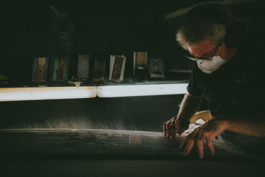 Shapers-Club- Une personne portant un équipement de protection travaille minutieusement sur une pièce de tissu à l'Univers du Shape, dans un atelier faiblement éclairé. -surfshop-surfboard