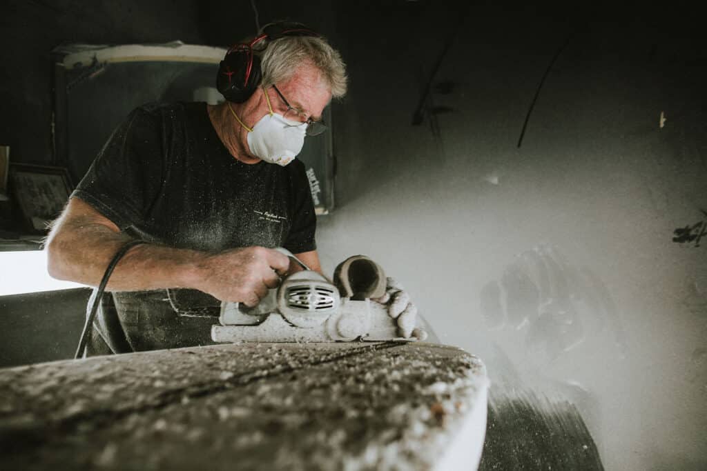 Shapers-Club- Une personne portant un masque anti-poussière et des lunettes de protection utilise une scie circulaire, coupant une dalle de pierre pour l'Univers du Shape et générant une quantité importante de poussière. -surfshop-surfboard