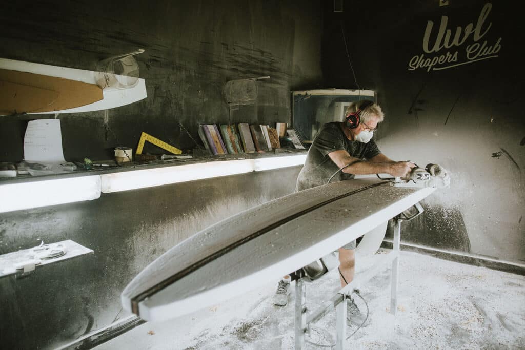 Shapers-Club- Un Maître Artisan des Planches de Surf portant un équipement de protection façonne une planche de surf dans un atelier poussiéreux. -surfshop-surfboard