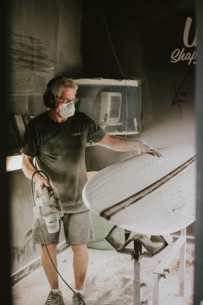 Shapers-Club- Une personne portant un équipement de protection utilise un outil électrique sur les planches de surf Roger Hinds dans un atelier, avec des particules de poussière visibles dans l'air. -surfshop-surfboard