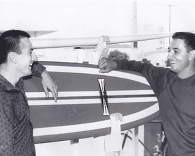Shapers-Club- Deux hommes souriants et appuyés sur une planche de surf Gordon & Smith. -surfshop-surfboard