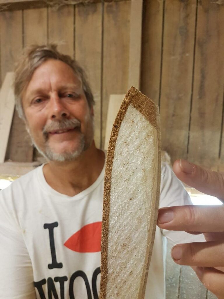 Shapers-Club- Homme souriant derrière une tranche de pain à la croûte inhabituellement épaisse, influencé par des influences culturelles. -surfshop-surfboard