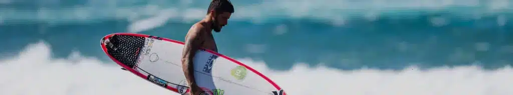 Shapers-Club- A surfer wearing a board with the best LEASH walking along the shore with waves in the background. -surfshop-surfboard