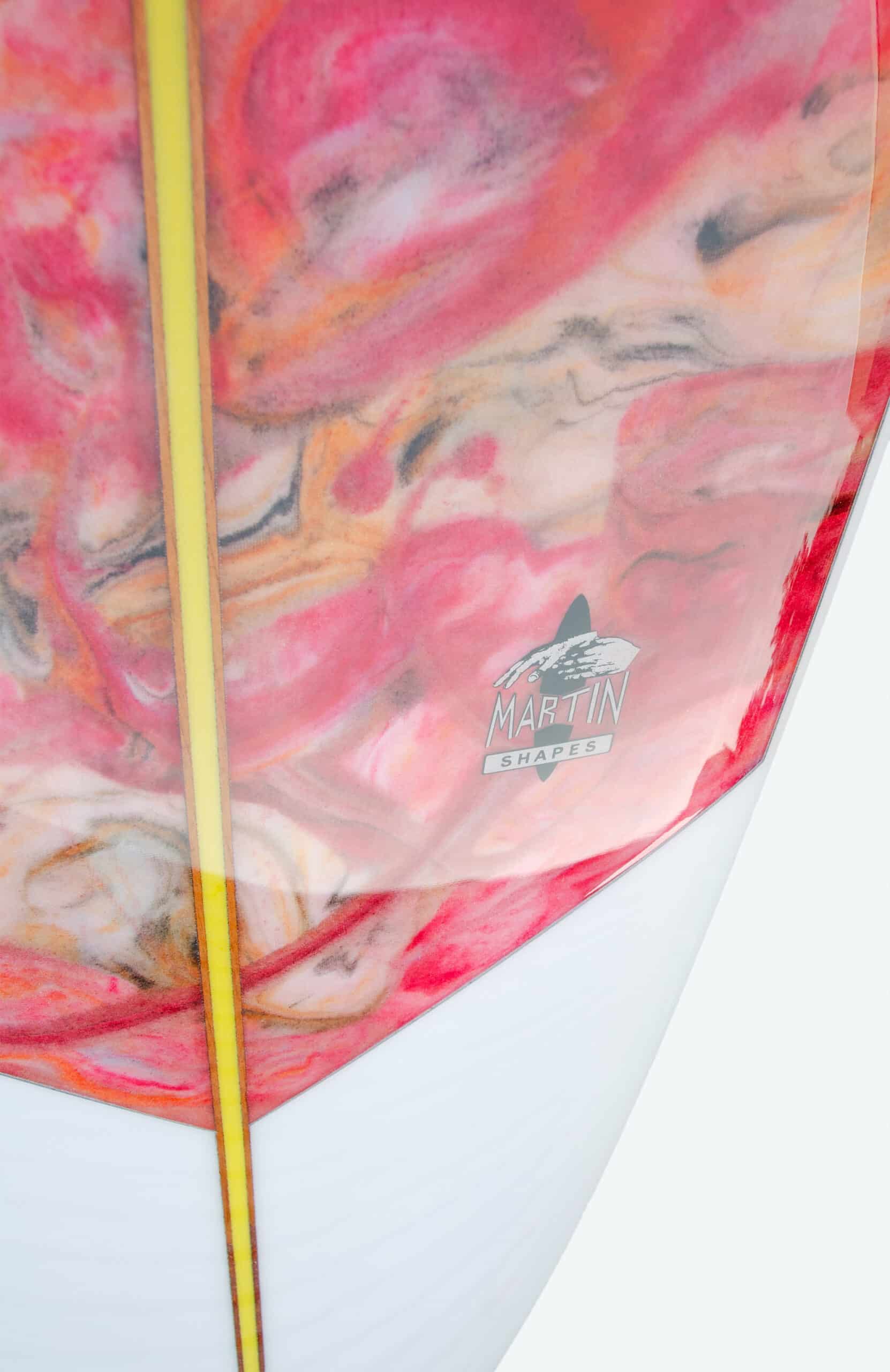 Shapers-Club- Gros plan de la planche de surf Martin Shape - Travelette, présentant un motif tourbillonnant vibrant en rose, orange et crème sur sa surface. Le dessous présente un petit logo « Martin Shapes » avec un graphisme de montagne stylisé. Cette planche de surf de 9'4" x 22' 3/4" x 3" présente également une bande jaune distinctive qui s'étend verticalement. -surfshop-surfboard