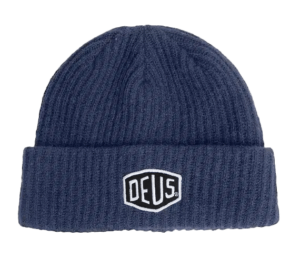 Shapers-Club- The Deus Beanie - SHIELD BEANIE is a blue knit beanie with a folded brim, adorned with a white and black embroidered patch that prominently displays "DEUS" in the center front. It features a ribbed texture for added style and comfort. -surfshop-surfboard