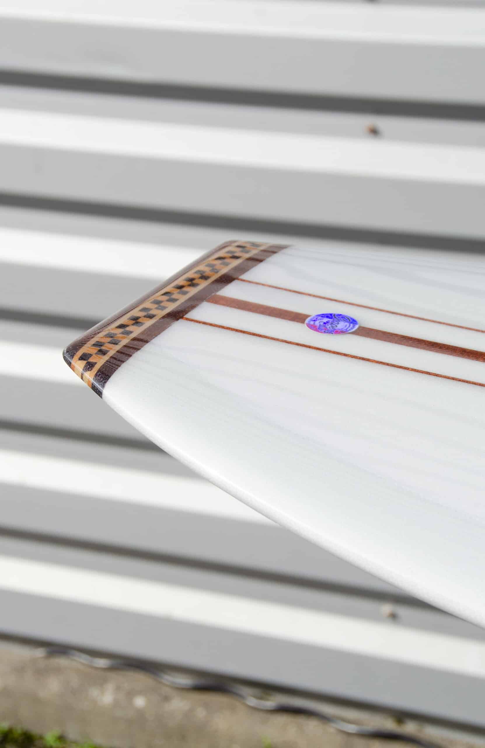 Shapers-Club- Primo piano della coda della tavola da surf «The Classic» di Roger Hinds, che misura 9'4" x 23" x 3'1/4", con strisce marroni e nere e un piccolo adesivo circolare blu. Lo sfondo è a strisce orizzontali grigie e bianche, probabilmente su una parete di metallo o di legno. -surfshop-surfboard