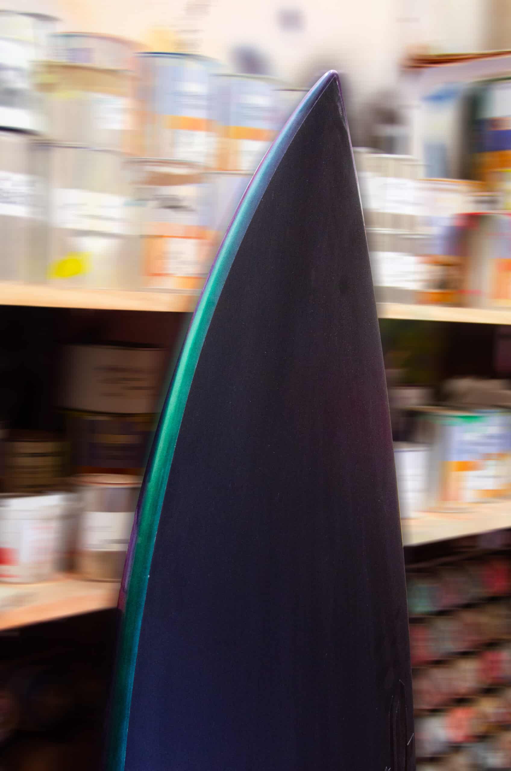 Shapers-Club- Un gros plan de la pointe d'une planche de surf Fire Arrow met en valeur son design élégant avec un dégradé de couleurs subtil. Cette planche noire de 5'9" x 19' 1/4" x 2' 1/2" est conçue pour la vitesse et l'agilité sur chaque vague, avec en arrière-plan des étagères remplies de canettes, de bocaux et de conteneurs divers. -surfshop-surfboard