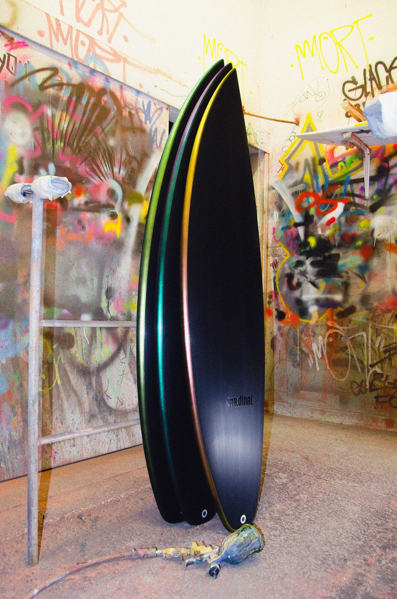 Shapers-Club- Trois planches de surf Cardinal Fire Arrow aux couleurs vives (5'9" x 19' 1/4" x 2' 1/2"), chacune avec une finition brillante, sont appuyées contre un mur dans un décor urbain orné de graffitis. Une sculpture abstraite se dresse à gauche, tandis que le sol est jonché de bombes de peinture et d'outils. Baignée de lumière dorée, cette scène rayonne d'énergie comme une flèche de feu saisissante dans le paysage urbain. -surfshop-surfboard
