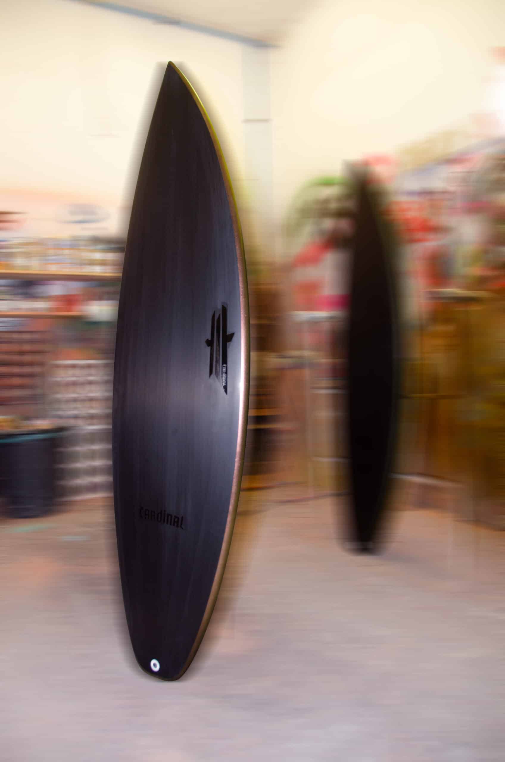 Shapers-Club- La planche de surf Fire Arrow 6'0" x 20" x 2' 5/8", avec sa finition noire brillante et ses fentes d'ailerons visibles, se tient debout à l'intérieur sur un arrière-plan flou. La pièce doucement éclairée comporte des étagères remplies d'objets colorés, amplifiant les dimensions frappantes de la planche. -surfshop-surfboard