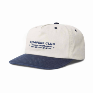 Shapers-Club- La CASQUETTES SHAPERS CLUB est une casquette de baseball beige avec un bord bleu marine. Elle comporte le texte "SHAPERS CLUB Custom surfboards" imprimé en bleu sur le devant, ainsi qu'un motif en zigzag bleu et un petit texte indiquant "1978" et "1981" de chaque côté du zigzag. -surfshop-surfboard