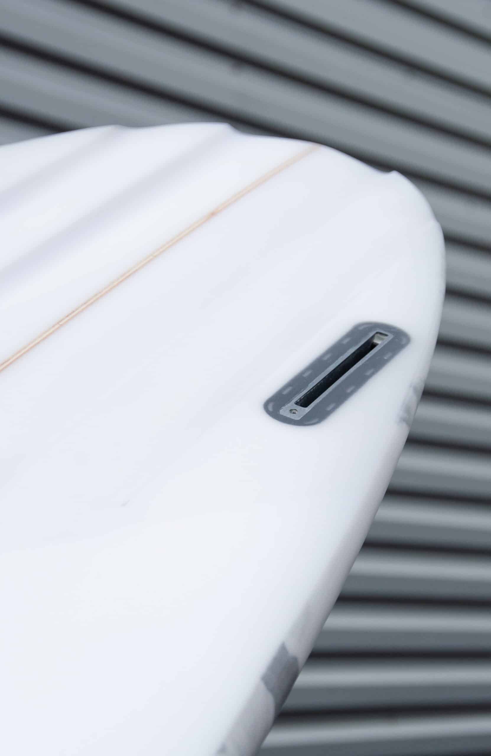 Shapers-Club- Gros plan de la queue d'une planche de surf blanche Aphex 6'10" x 20' 3/4" x 2' 3/4", mettant en valeur son boîtier à aileron unique, avec un revêtement en métal ondulé gris en arrière-plan. -surfshop-surfboard