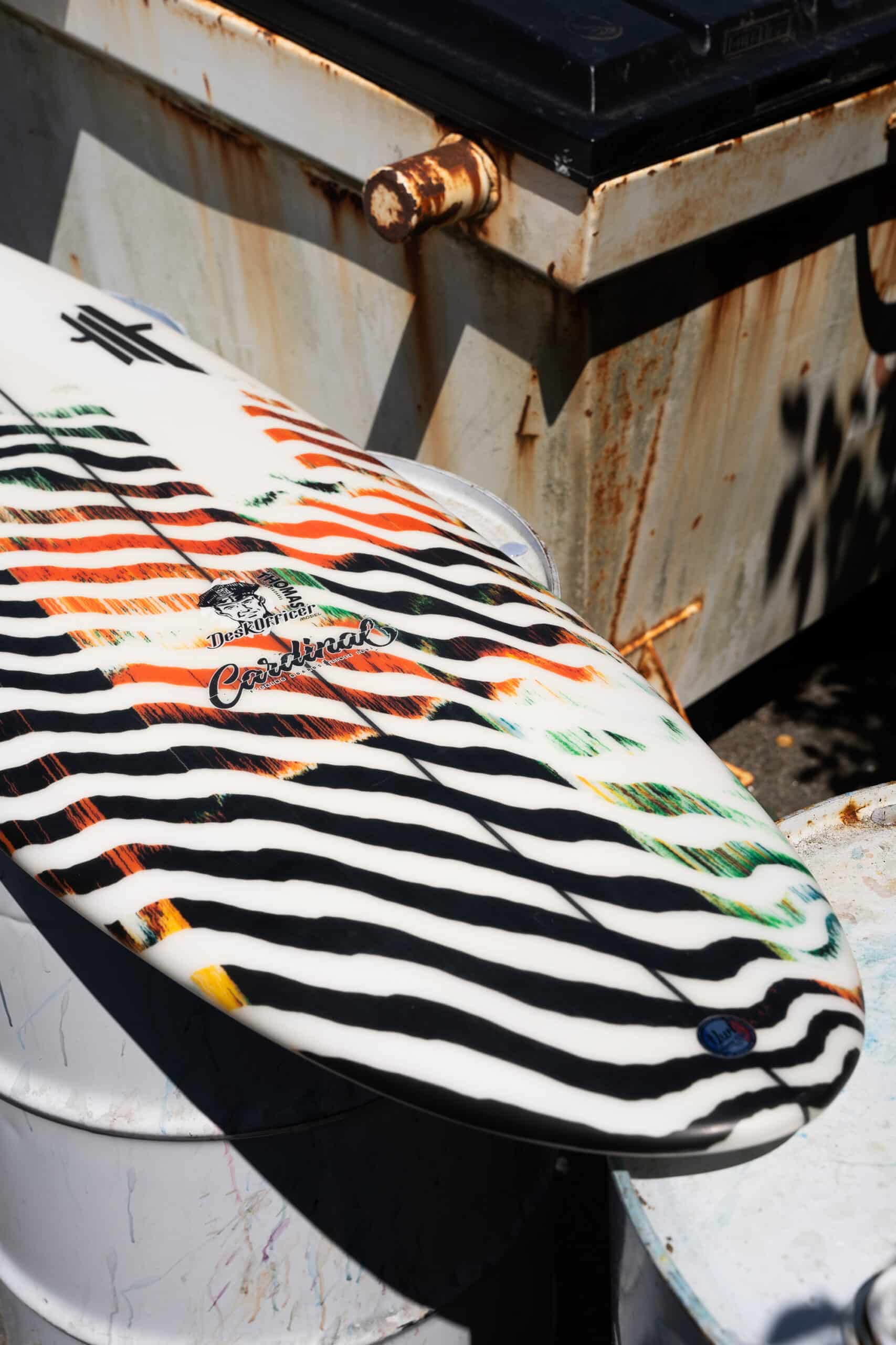 Shapers-Club- The Desk Officer surfboard, 5'7" x 19" x 2' 3/8", features bold black and white zebra stripes with touches of orange and green, leaning against rusty metal barrels. It perfectly blends industrial and sporty atmospheres, creating a scene even interior office officers could dream of amidst the paperwork. -surfshop-surfboard