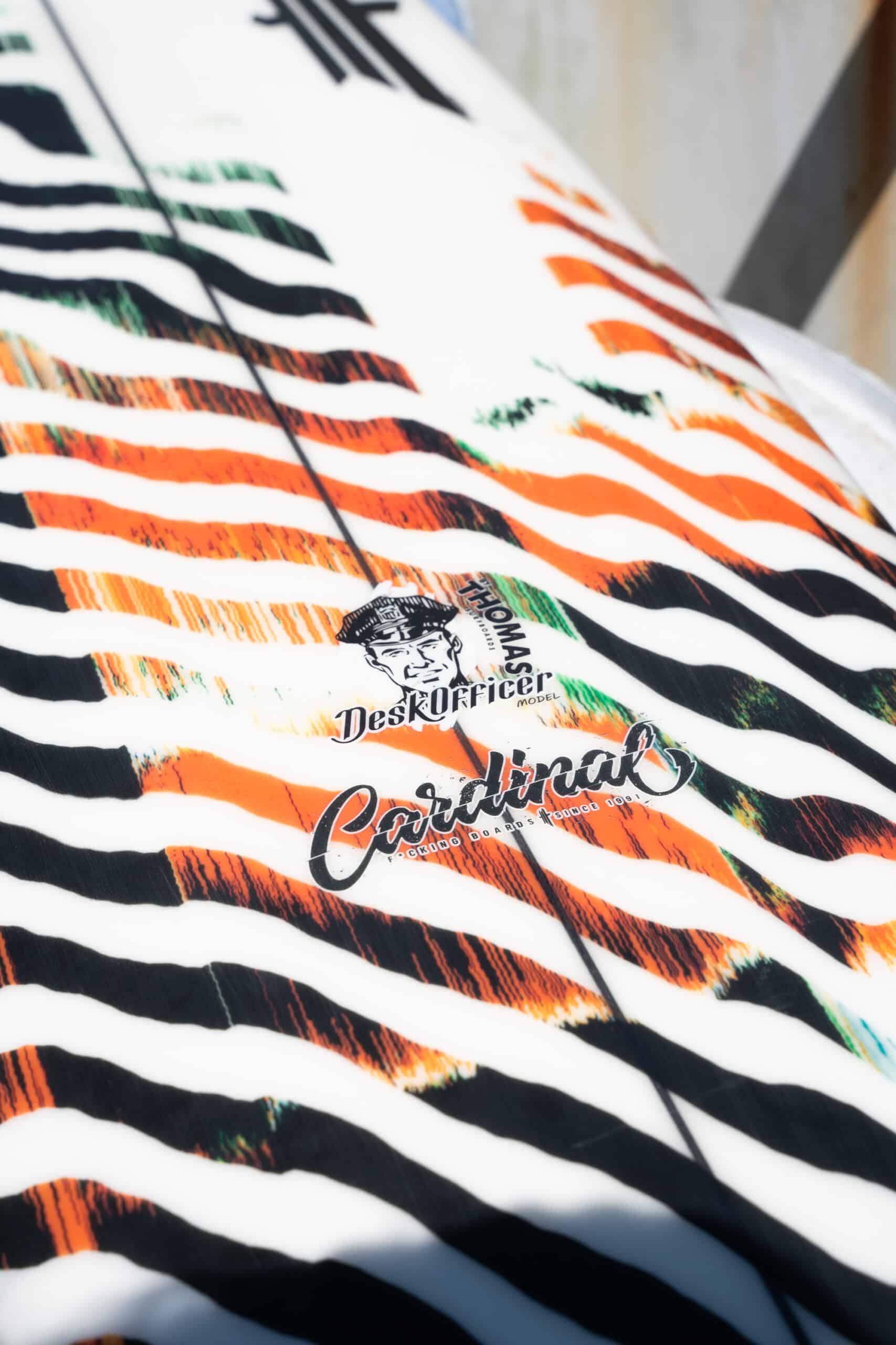 Shapers-Club- Close-up of a surfboard with bright orange, green and black stripes. It features "The Mason Desk Officer" and "Caribbean" logos, with sleek dimensions of 5'7" x 19" x 2' 3/8", enhancing its dynamic appeal. -surfshop-surfboard