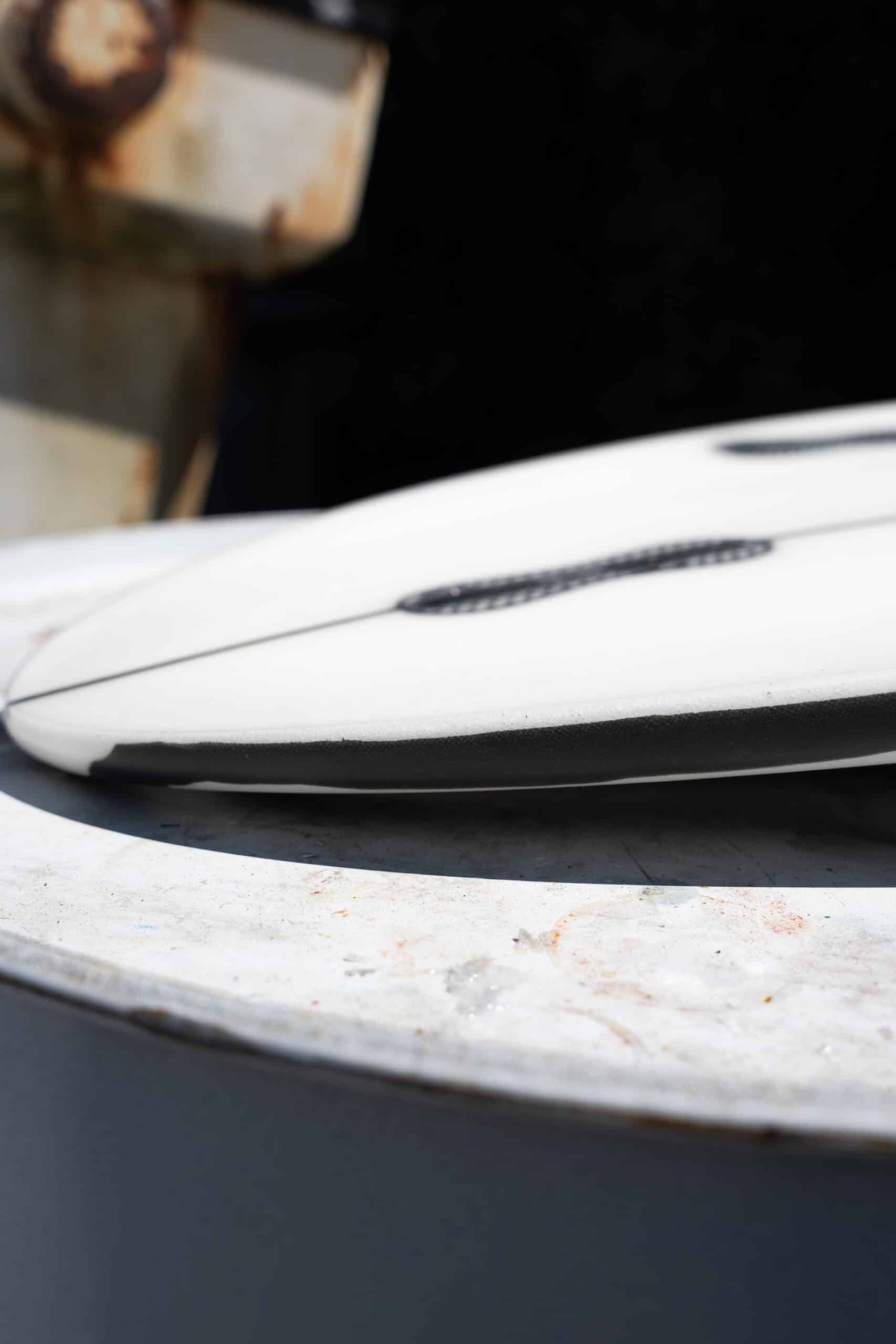 Shapers-Club- Close-up of the white and black surface of the Desk Officer surfboard, set on concrete, echoing the lines of the office furniture, with a rusted metal background. Its dimensions of 5'7" x 19" x 2'3/8" create unique visible grooves. -surfshop-surfboard