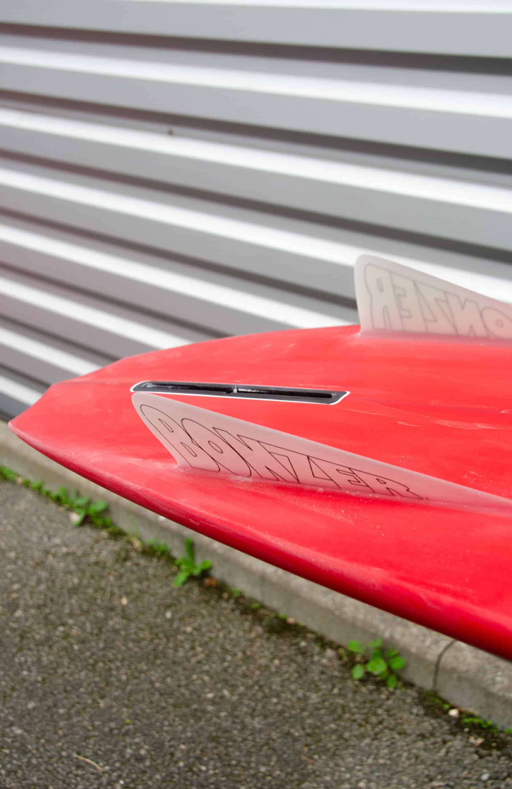 Shapers-Club- Gros plan de la queue de la planche de surf Russ Short en rouge avec un aileron et « BONZER » affichés, mesurant 6'2" x 19' 1/2" x 2' 1/2", sur du gravier près d'un mur en tôle ondulée. -surfshop-surfboard